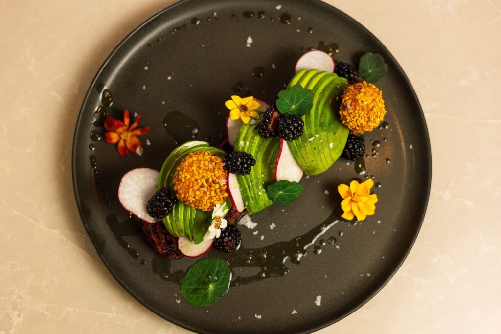 Plated course with avocado, radish, blackberries and edible flowers on a dark plate