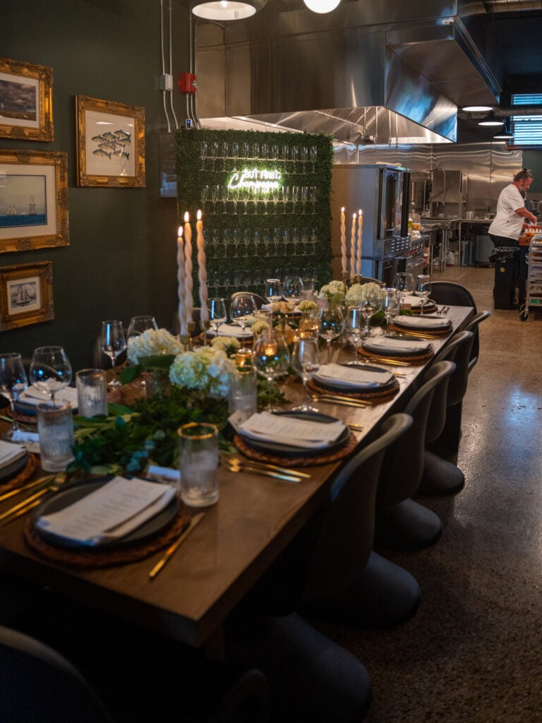 Long dinner table set with candles, greenery and glassware in front of an open kitchen