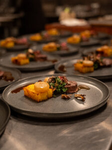 Plated course of sliced beef with polenta and greens, lined up for service